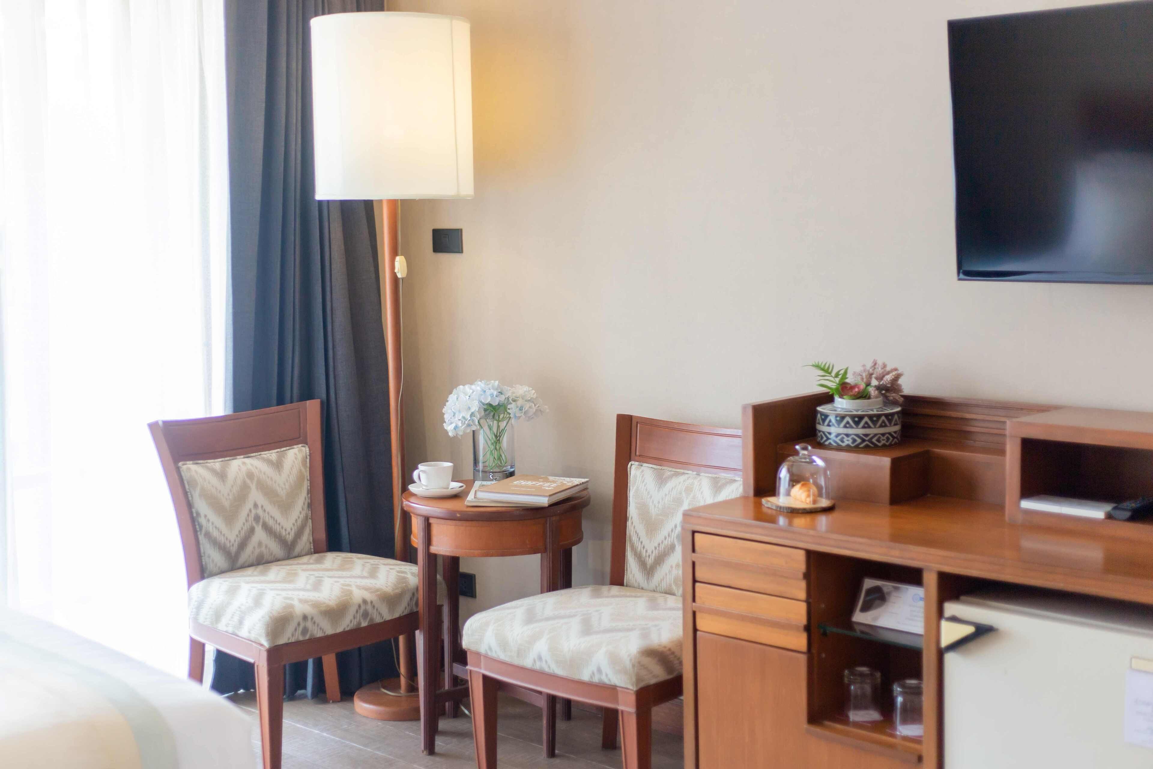 A hotel room with a wooden desk, two chairs, a lamp, a television, and a small table with a vase and a book.