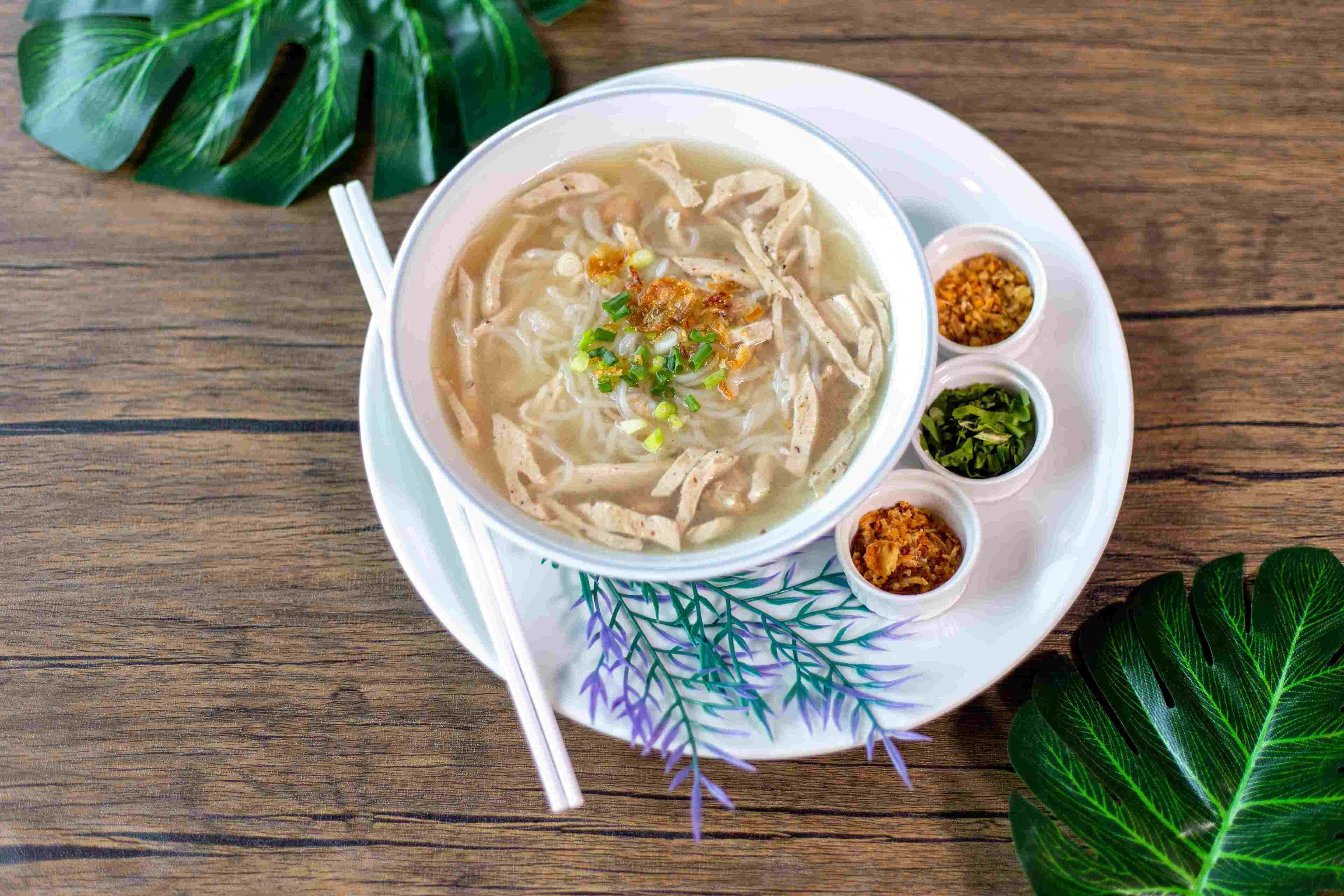 Un plato de sopa con fideos y pollo, acompañado de recipientes pequeños con condimentos sobre un plato blanco con diseño de hojas decorativas.