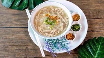 Un plato de sopa con fideos y pollo, acompañado de recipientes pequeños con condimentos sobre un plato blanco con diseño de hojas decorativas.