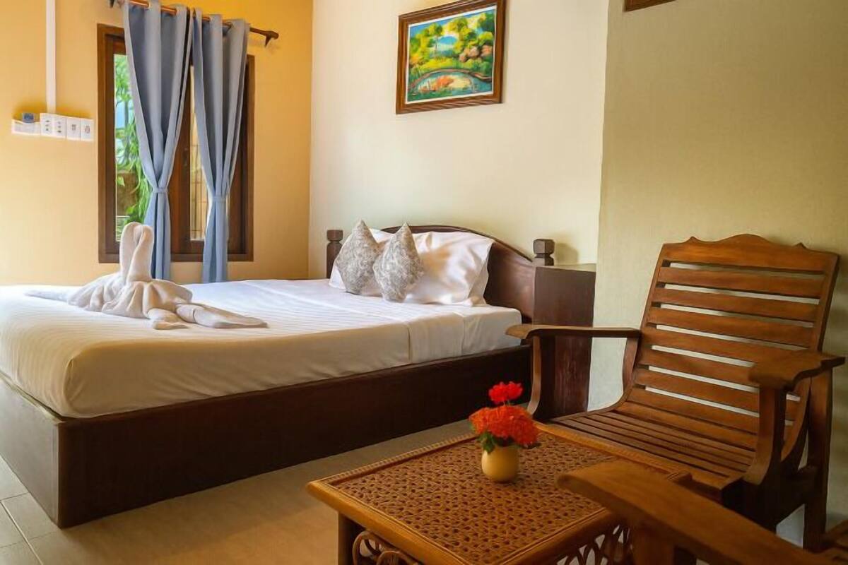 A hotel room with a bed, a wooden chair, a small table with a vase of flowers, and a painting on the wall.