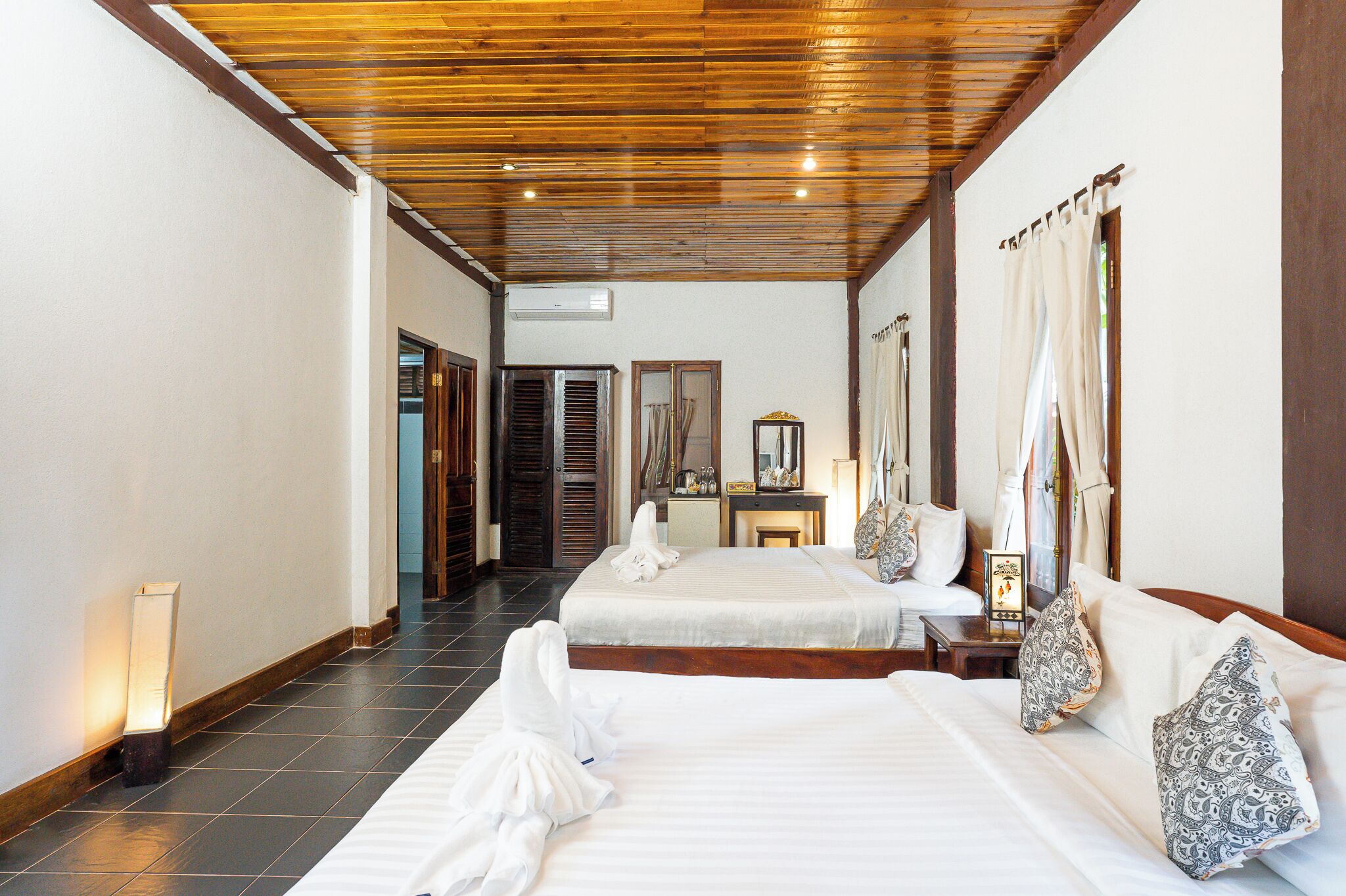 Habitación de hotel con dos camas, techo de madera, una mesita de noche, un espejo y una ventana con cortinas.