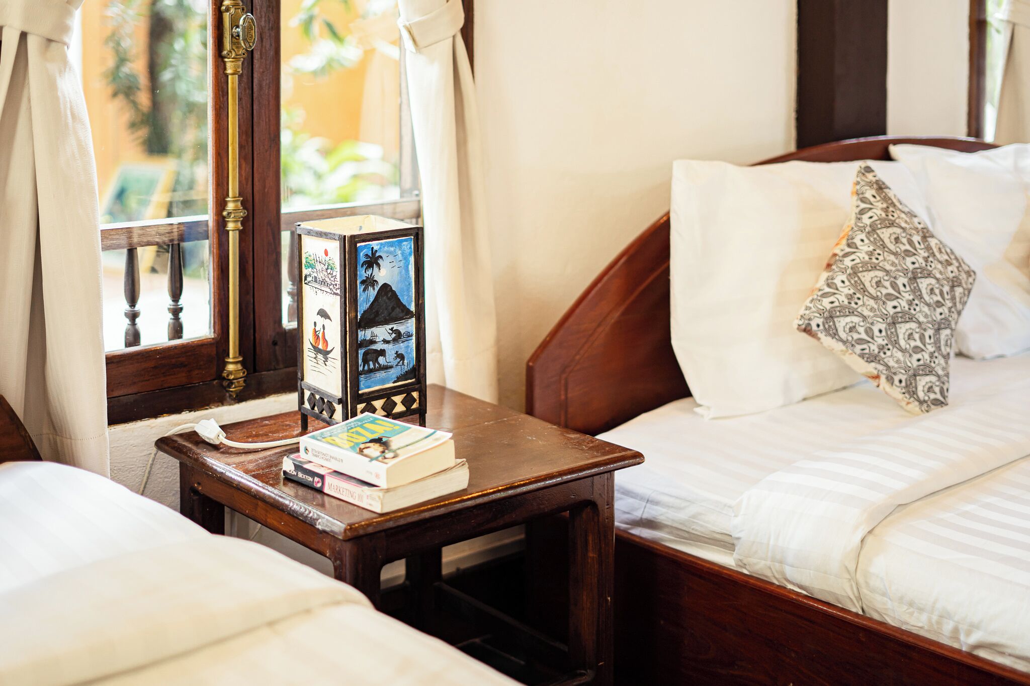 A neatly made bed with white linens and a patterned pillow, a bedside table with books and a framed picture, and a window with curtains.