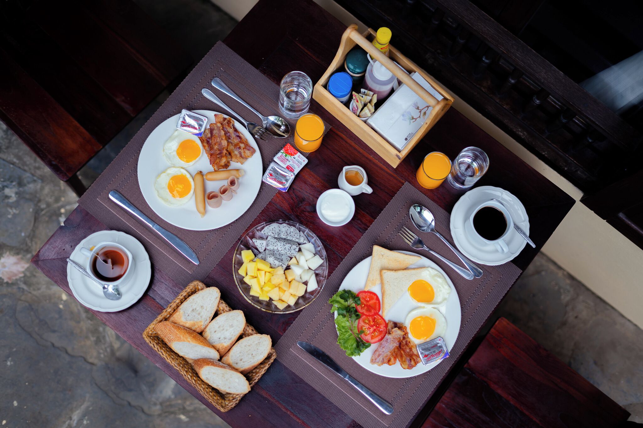 A breakfast table with plates of eggs, bacon, toast, and fruit. There are also condiments, a cup of coffee, and a basket of bread.