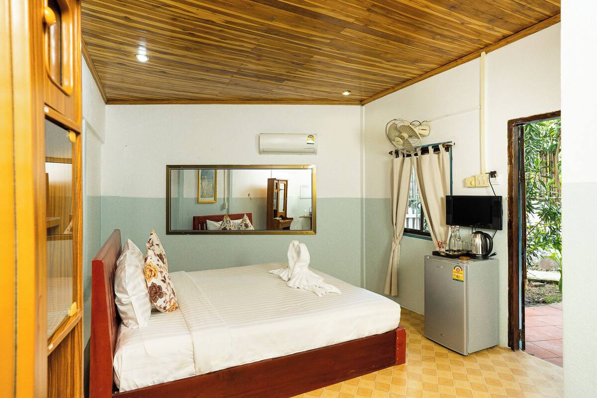 A hotel room with a wooden ceiling, a bed with white linens, a small refrigerator, a television, and a window with curtains.