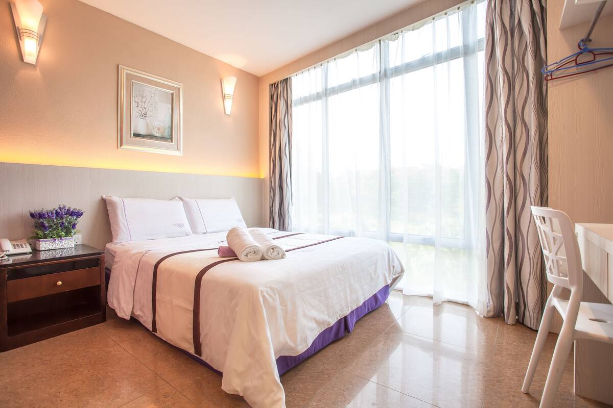 A hotel room with a bed, bedside table, lamp, and a view of the outdoors through the window.