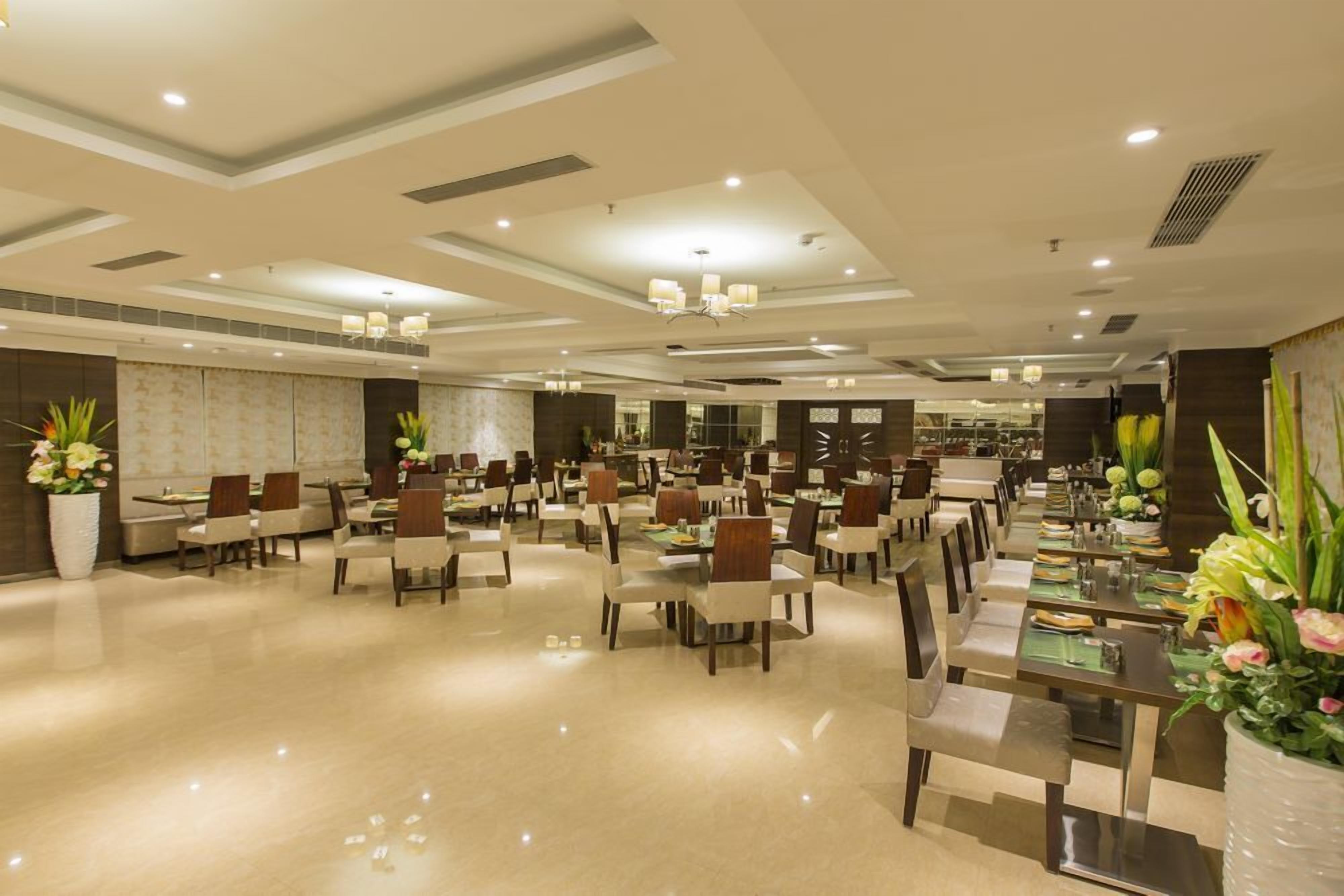 Ampla sala de jantar de hotel, com várias mesas e cadeiras, bem iluminada por luminárias de teto e decorada com plantas em vasos.