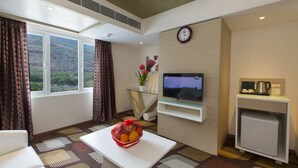 Habitación de hotel con un ventanal grande, un televisor de pantalla plana, un refrigerador pequeño, una cafetera y una cesta de frutas sobre una mesa blanca.