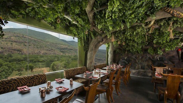 Un restaurante con vistas a un paisaje montañoso, muebles de madera y un gran árbol con follaje colgante.