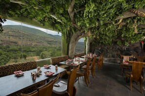 A restaurant with a view of a mountainous landscape, wooden furniture, and a large tree with hanging foliage. - Pai Viceroy (Tirupati)