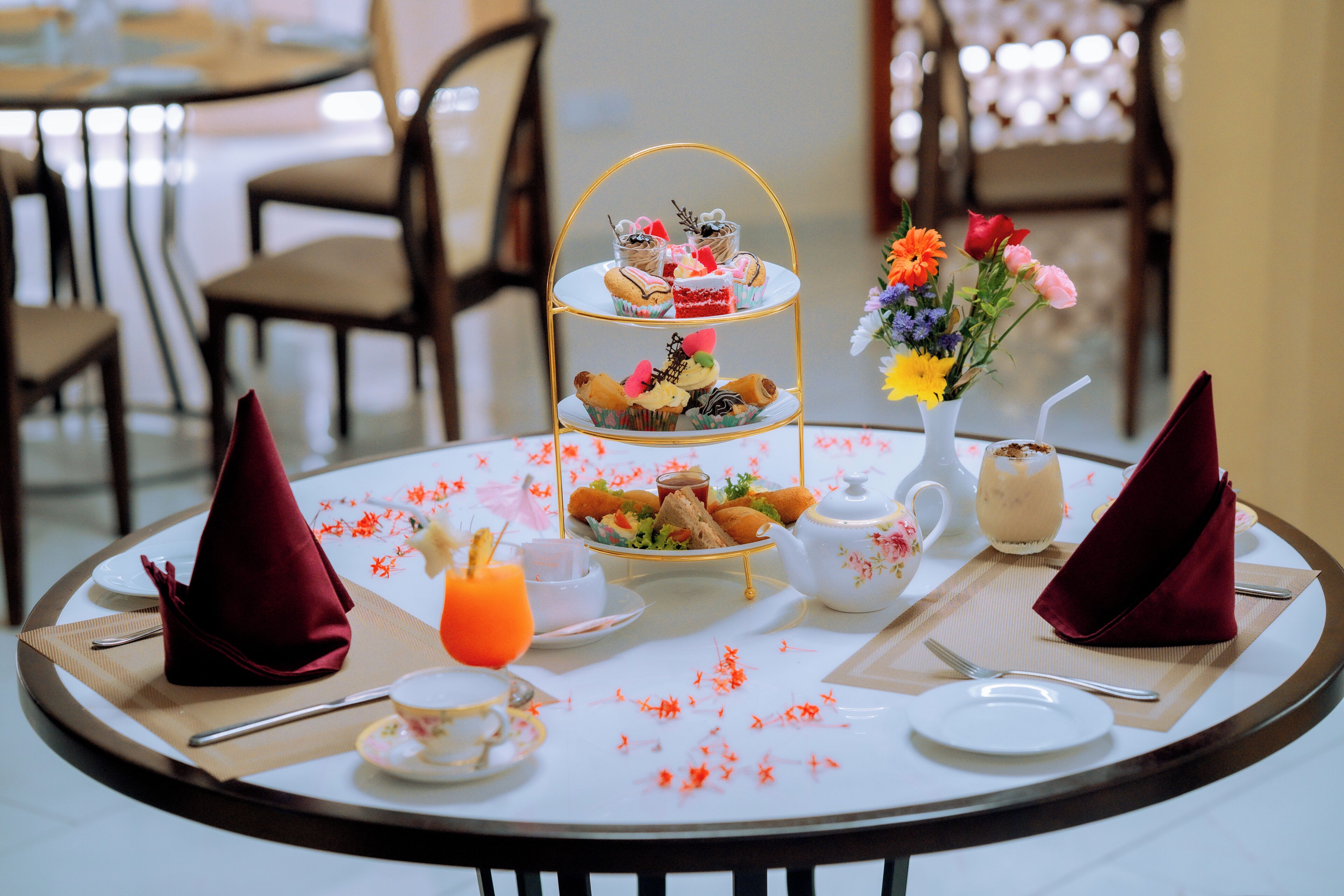 A round dining table with a tiered dessert stand, a vase of flowers, a teapot, and a glass of orange juice.