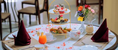A round dining table with a tiered dessert stand, a vase of flowers, a teapot, and a glass of orange juice.