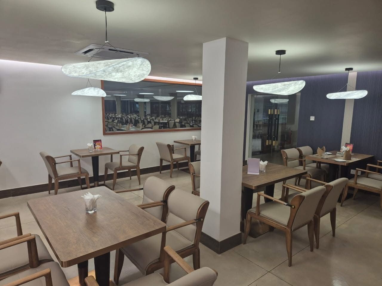Un restaurant avec des tables et des chaises en bois, des luminaires suspendus et un grand miroir au mur.