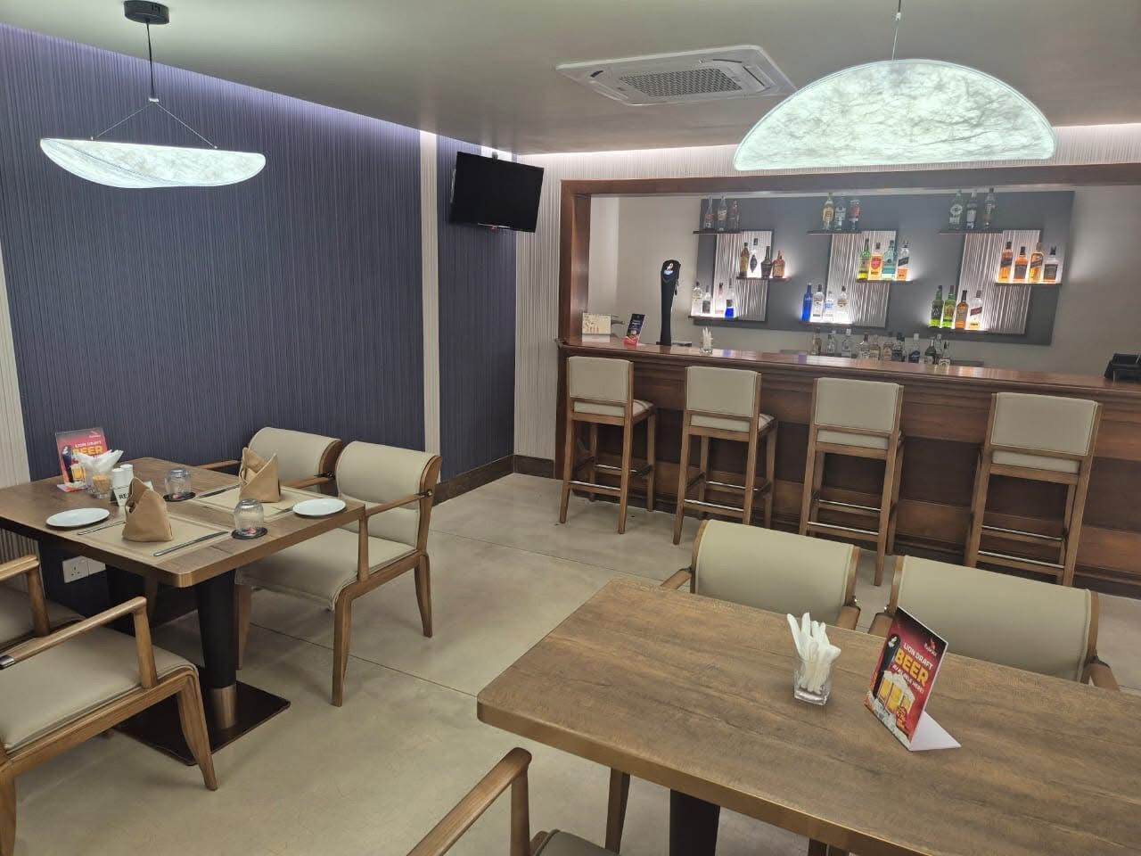 A restaurant with wooden tables and chairs, a bar area with bottles, and a television on the wall.