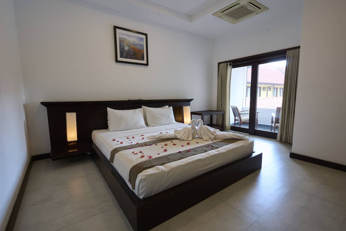 A hotel room with a large bed, a nightstand, a framed picture on the wall, and a view of the outside through a large window.