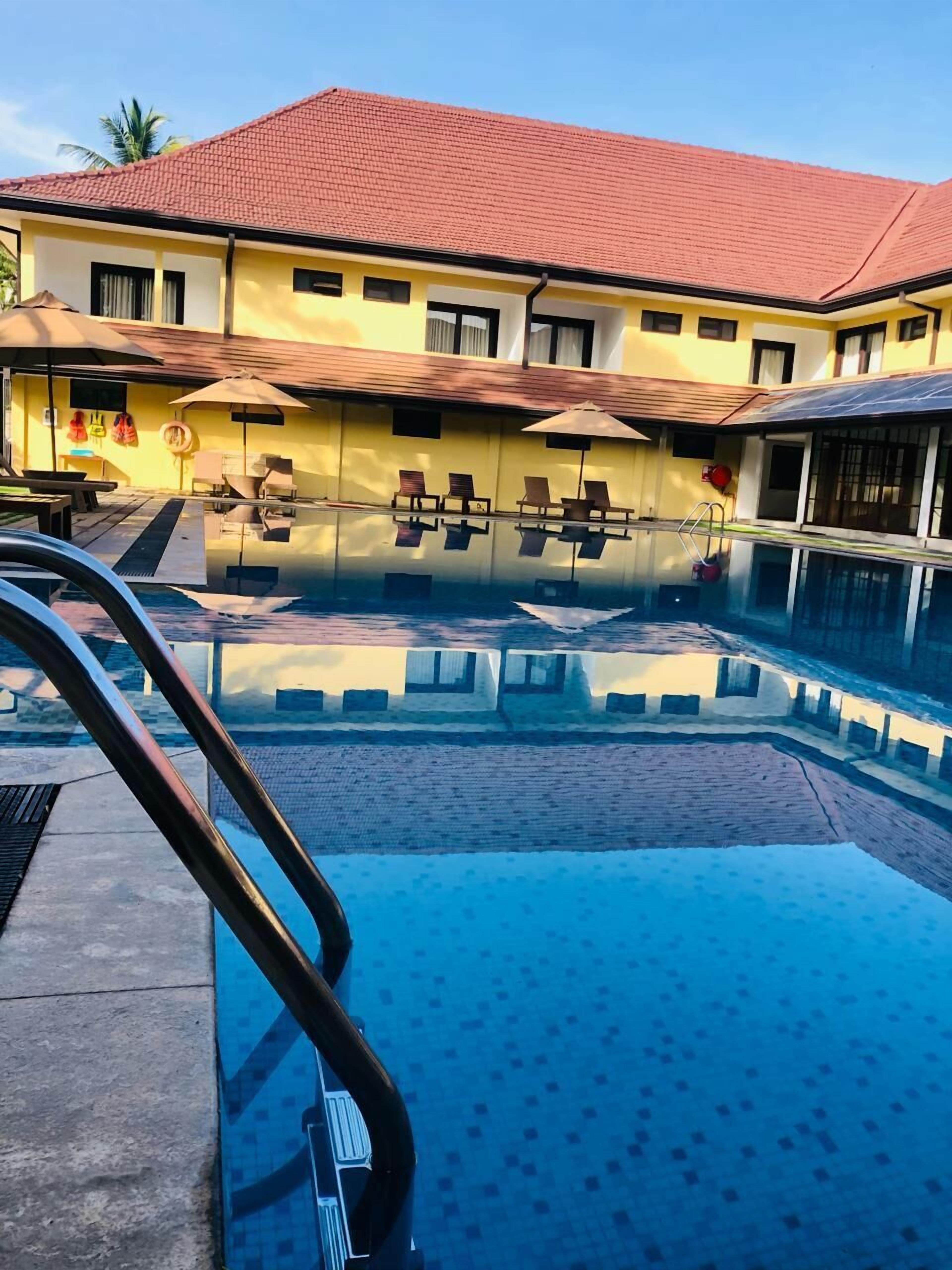 A swimming pool with a clear blue water surface, a red-tiled roof, and a yellow building with multiple windows and a balcony.