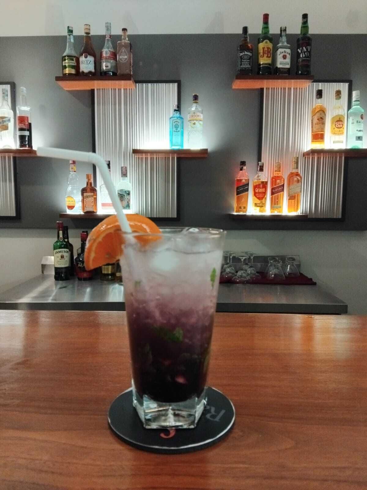 A bar area with shelves of liquor bottles, a glass with a drink and an orange slice, and a coaster with a logo.