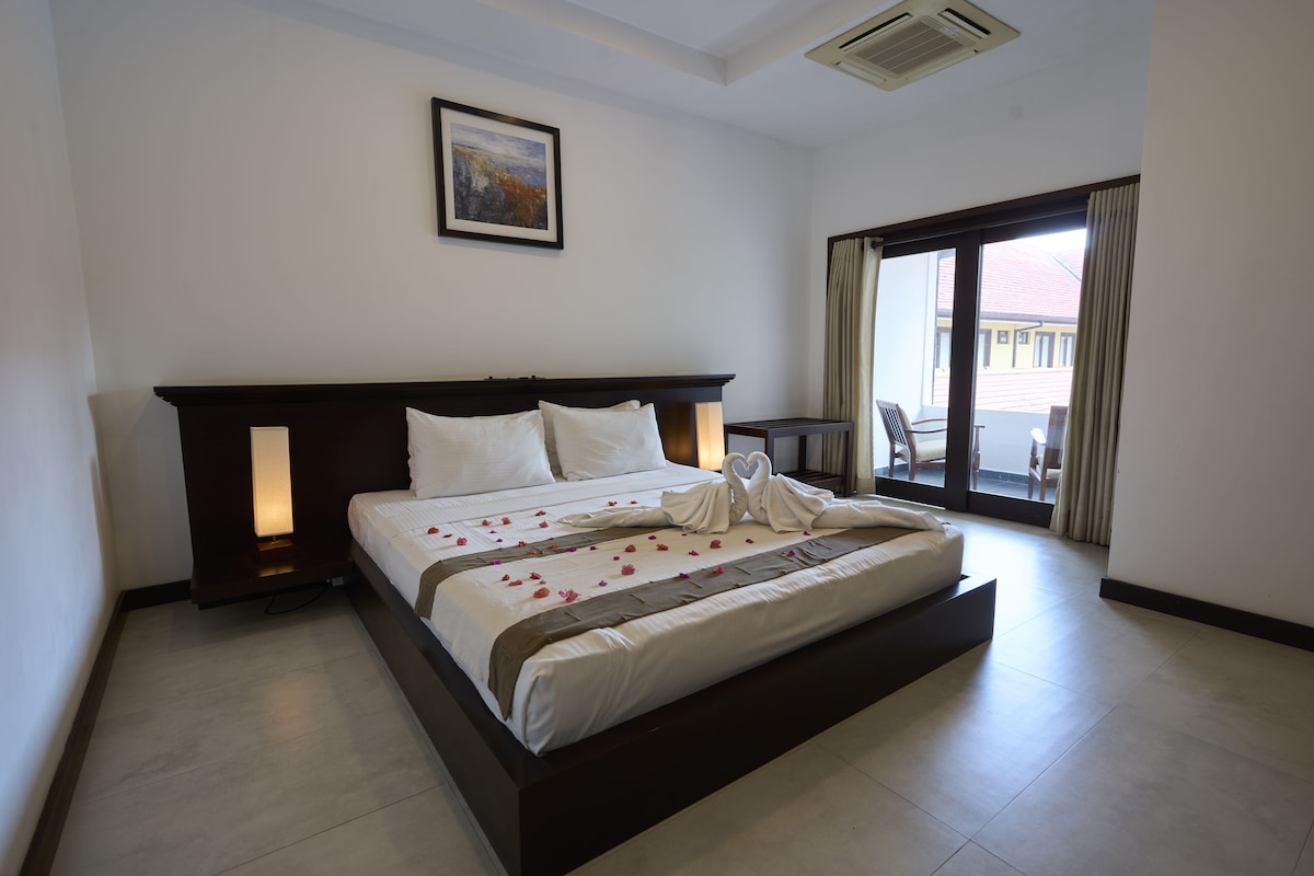 Habitación de hotel con una cama grande, una mesita de noche, un cuadro enmarcado en la pared y vistas al exterior a través de un amplio ventanal.