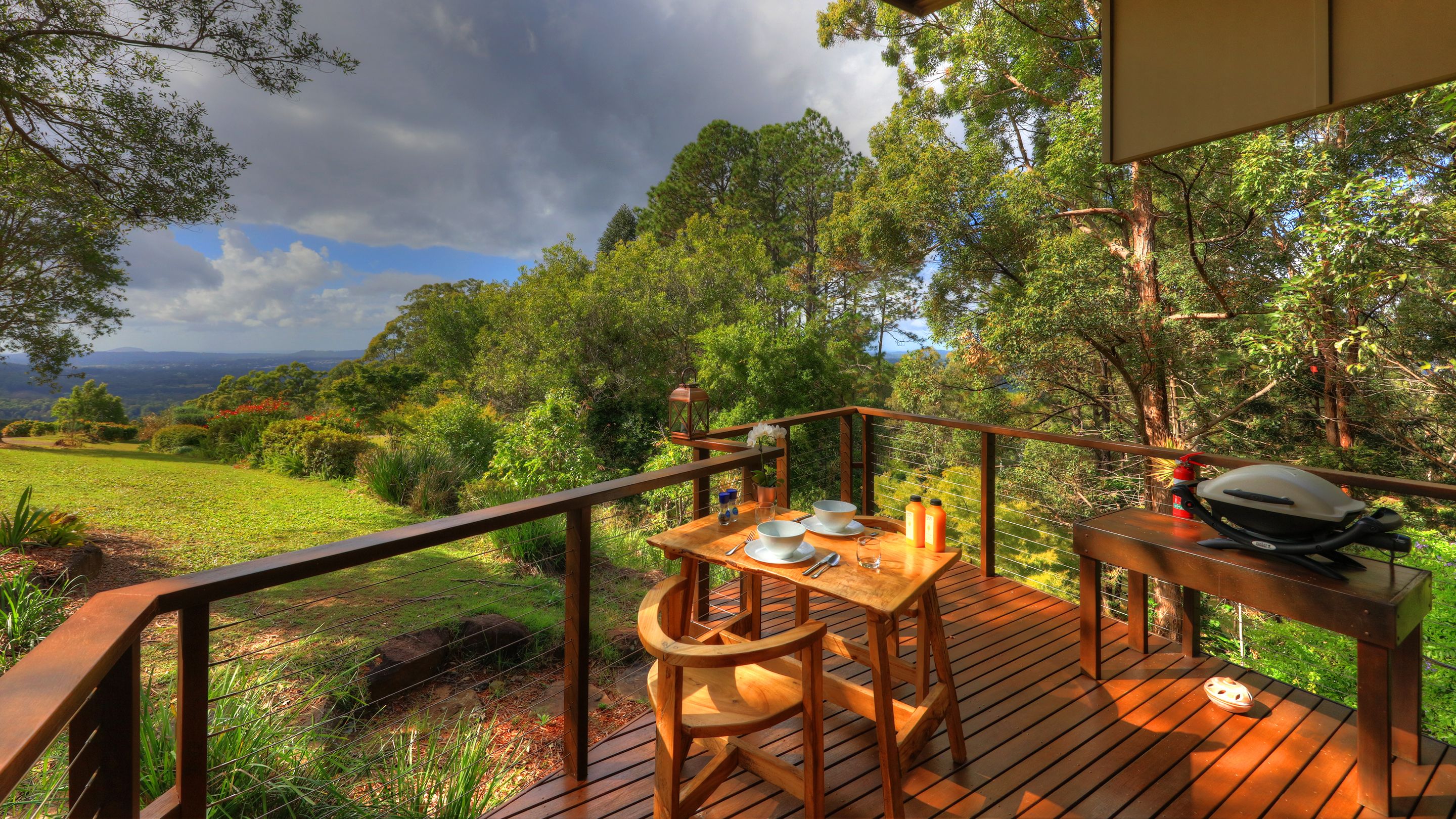 A wooden deck with a table set for dining, overlooking a lush landscape with trees and a distant view of hills.