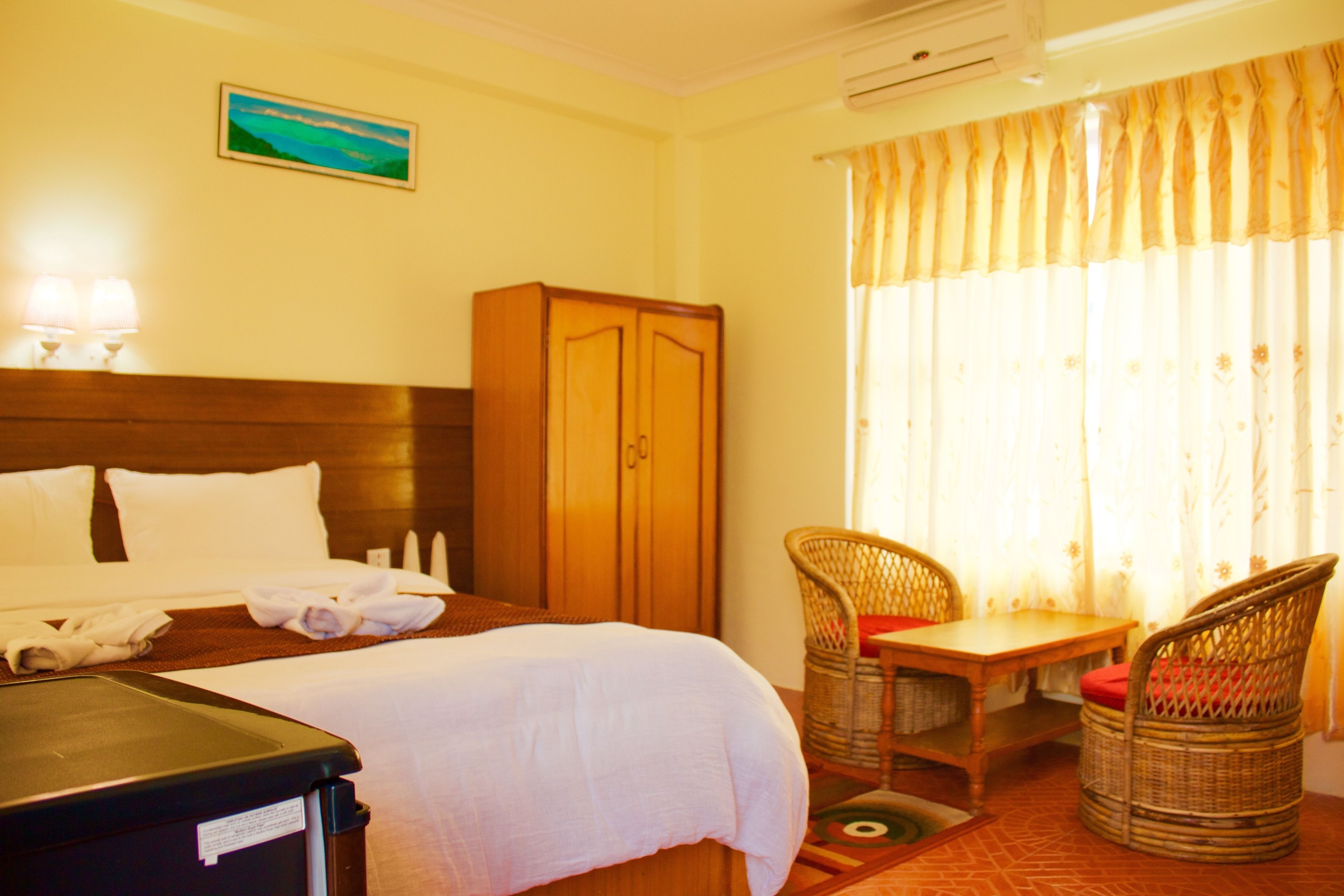 A hotel room with a bed, a wooden wardrobe, a desk with a chair, and a window with curtains.