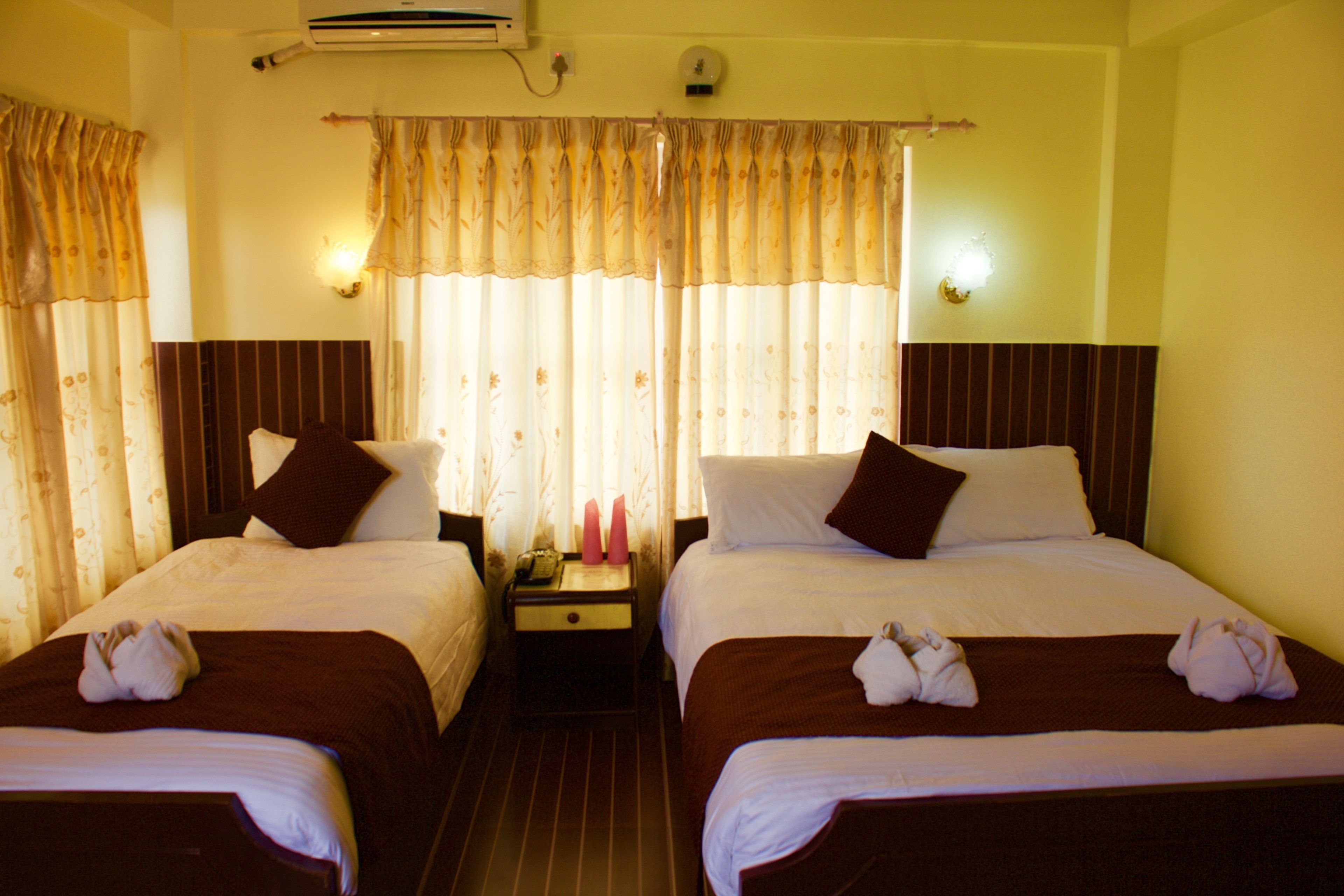 Habitación de hotel con dos camas, cada una con una colcha marrón y almohadas blancas. Entre las camas, un mesita de noche con una lámpara y un teléfono.