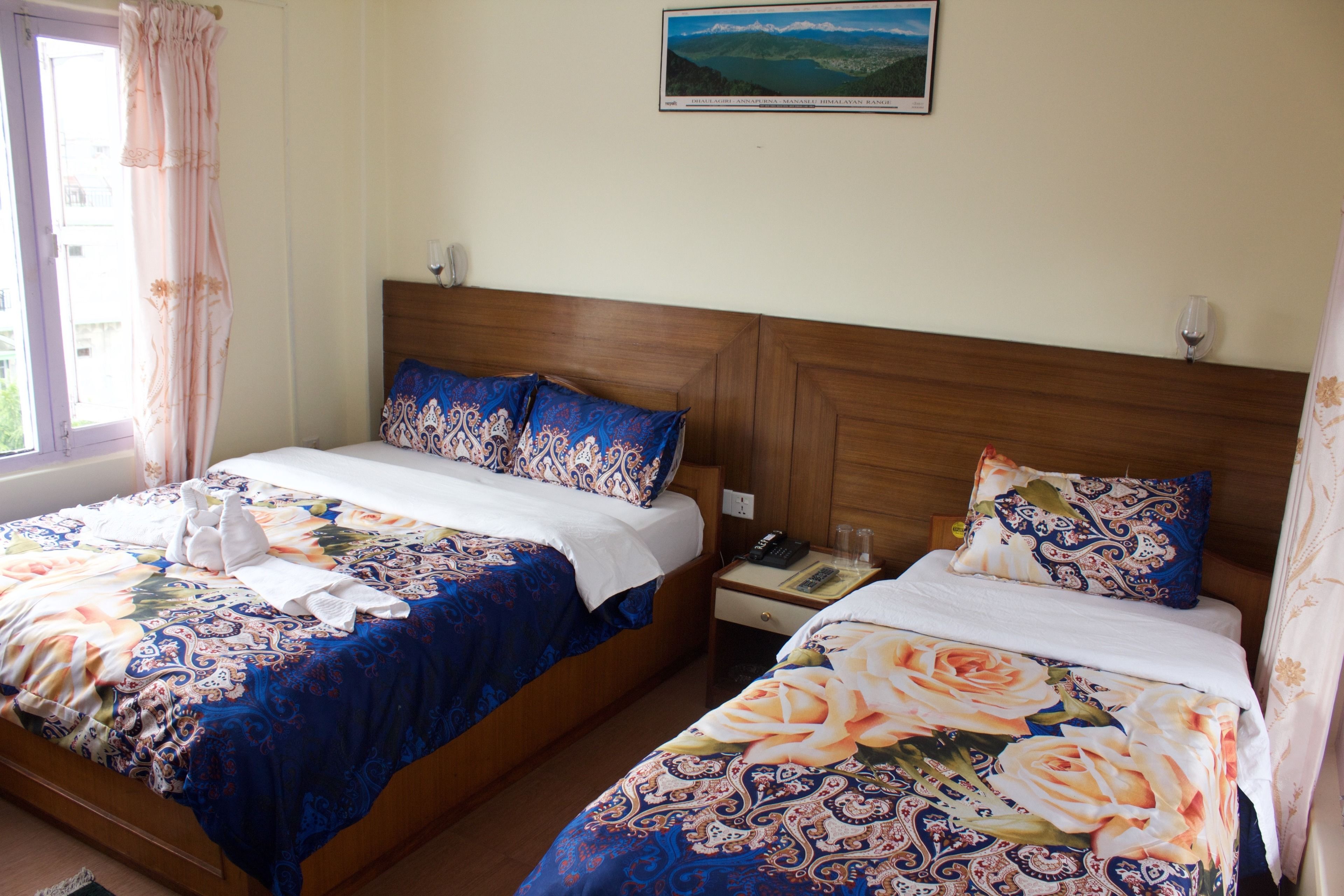 A double bed with floral bedding and a wooden headboard, a bedside table with a phone and glass, and a window with curtains.