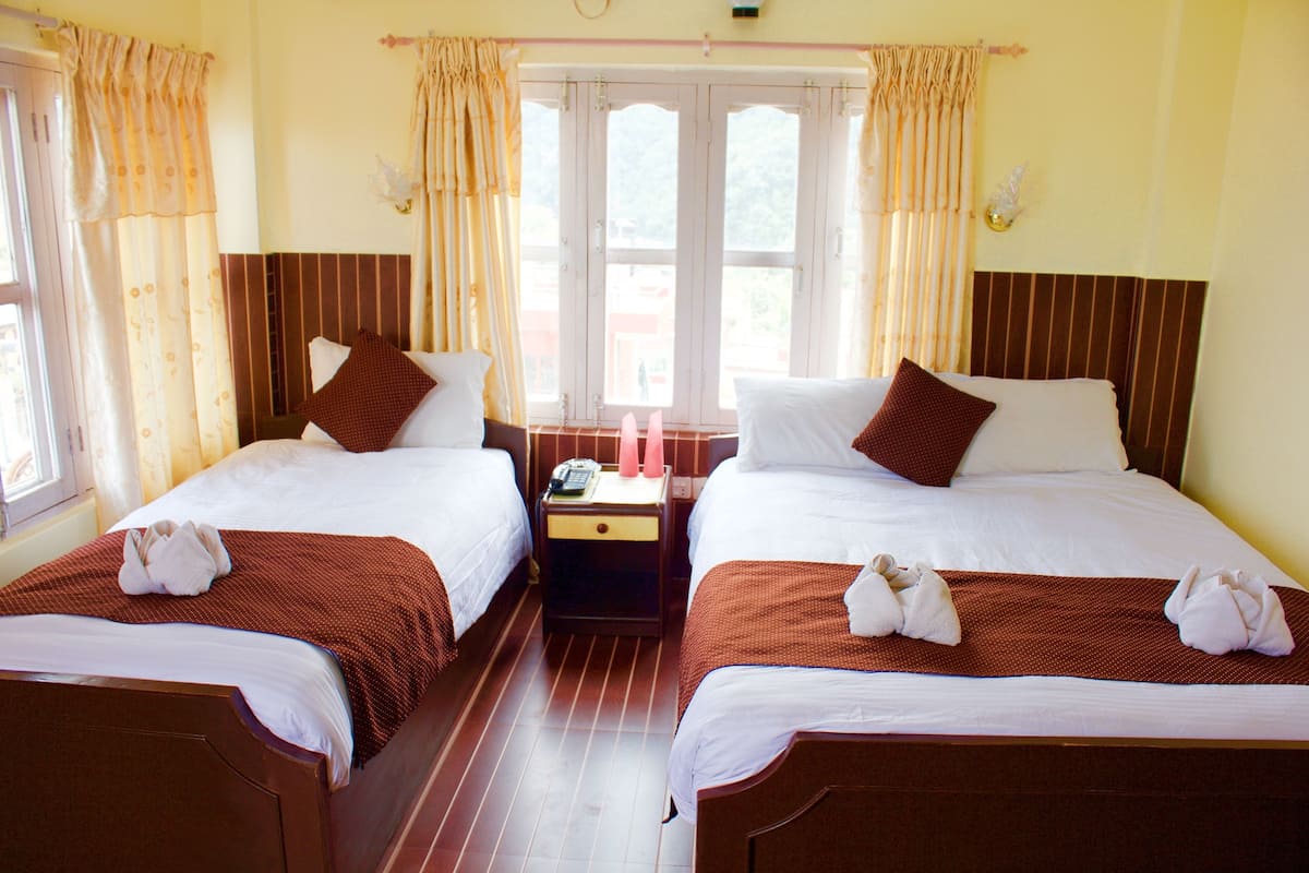 Une chambre d’hôtel avec deux lits, chacun recouvert d’une couvre-lit marron et agrémenté d’oreillers blancs. Une table de chevet avec un téléphone et une petite lampe se trouve entre les deux lits. La chambre est dotée d’une fenêtre avec des rideaux et offre une vue sur l’extérieur.