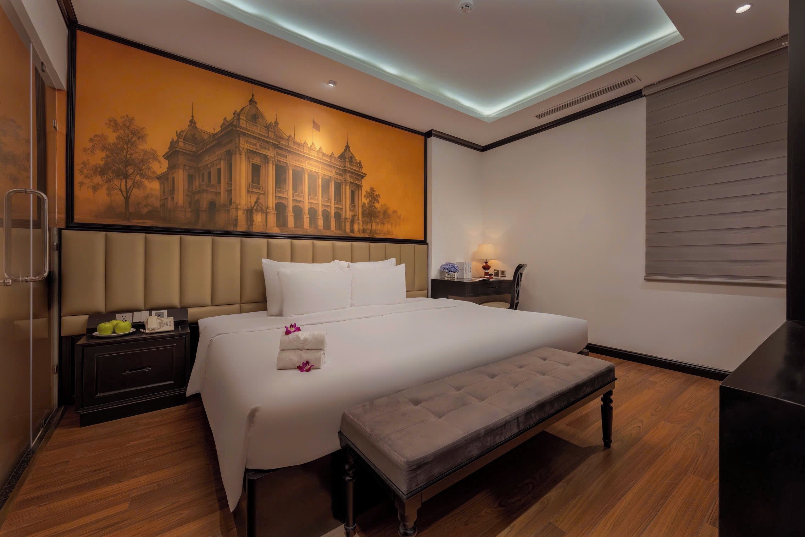 A hotel room with a large bed, a bench, a nightstand, a desk, and a wall-mounted lamp.