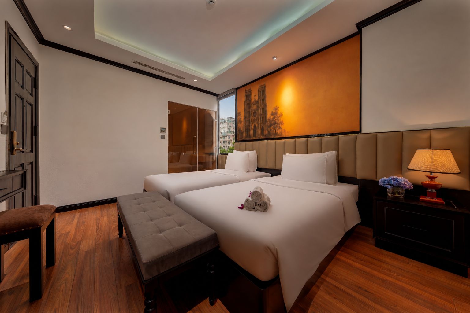 Een hotelkamer met twee bedden, een bankje, een nachtkastje met een lamp en een groot raam.