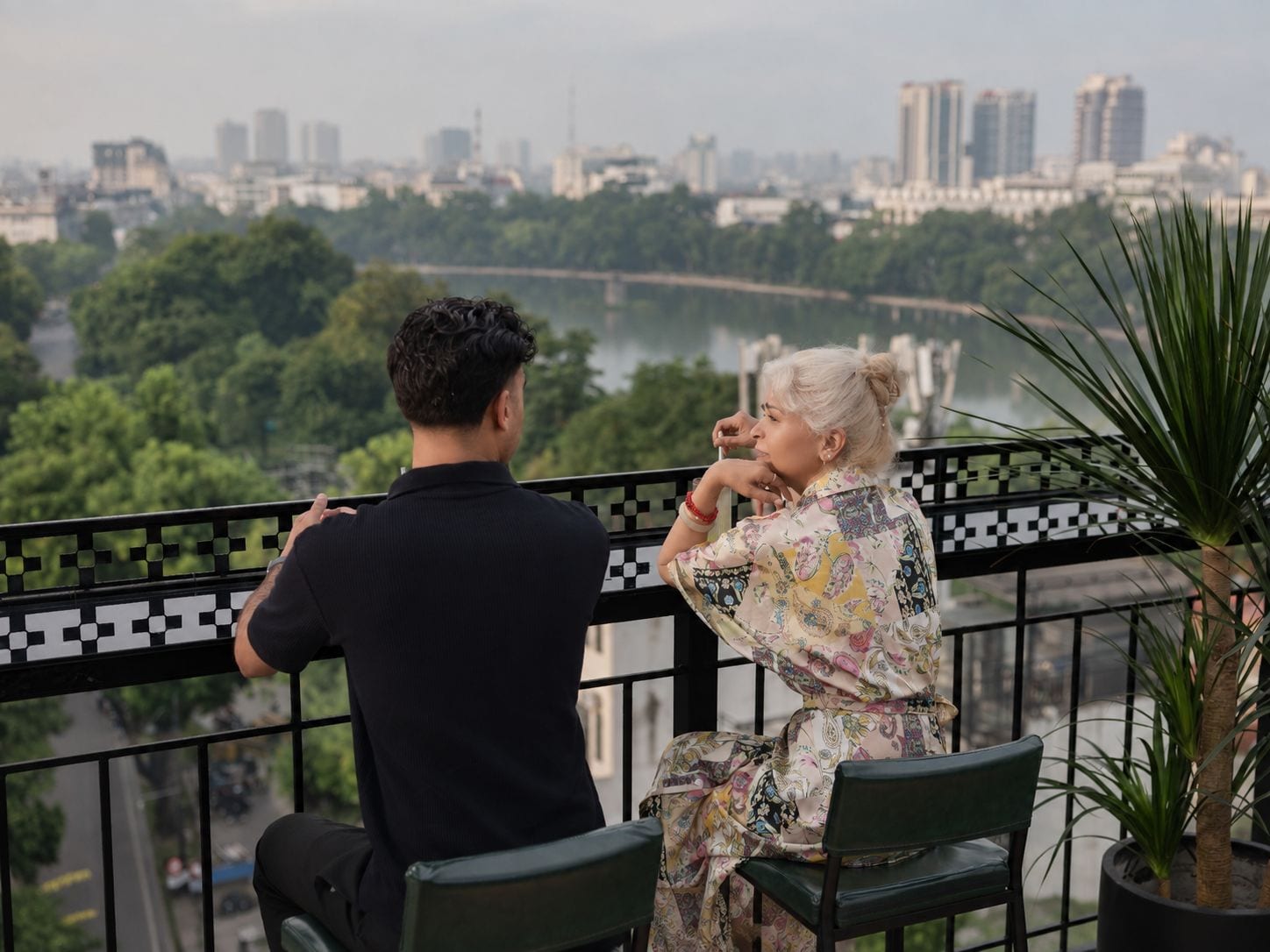 Two people seated on a balcony with a cityscape view.