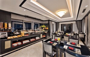 A modern hotel dining area with a buffet, seating, and a view of the city at night.