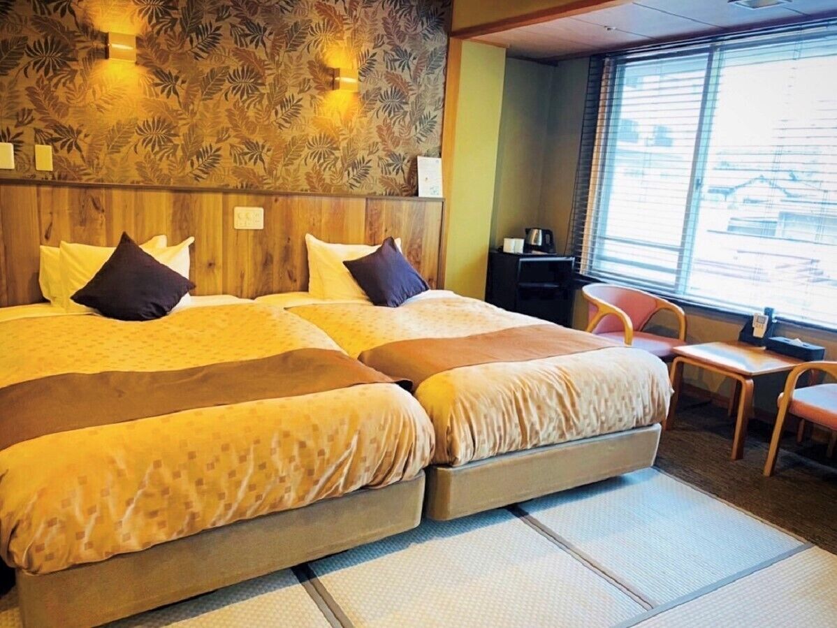 A room with a large bed, a small table, a chair, and a window with blinds.