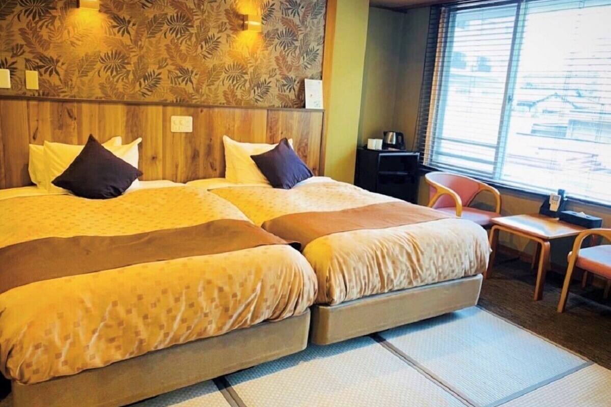 A room with a large bed, a small table, a chair, and a window with blinds.