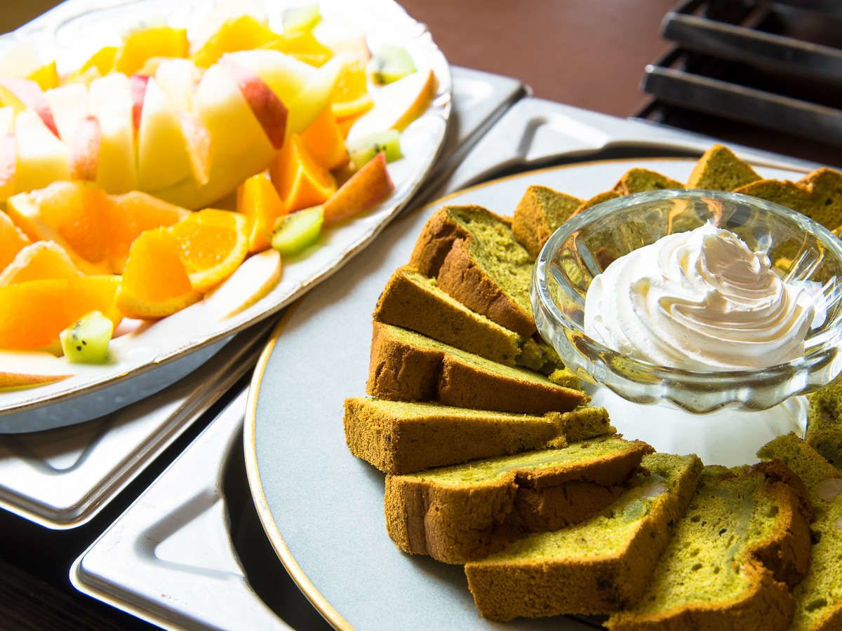 A plate of sliced green cake with a glass of white sauce, and a plate of fruit on the table.