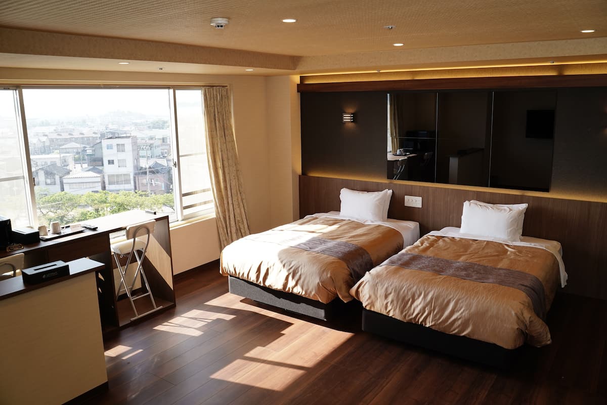 A hotel room with two beds, a desk, and a large window offering a city view.