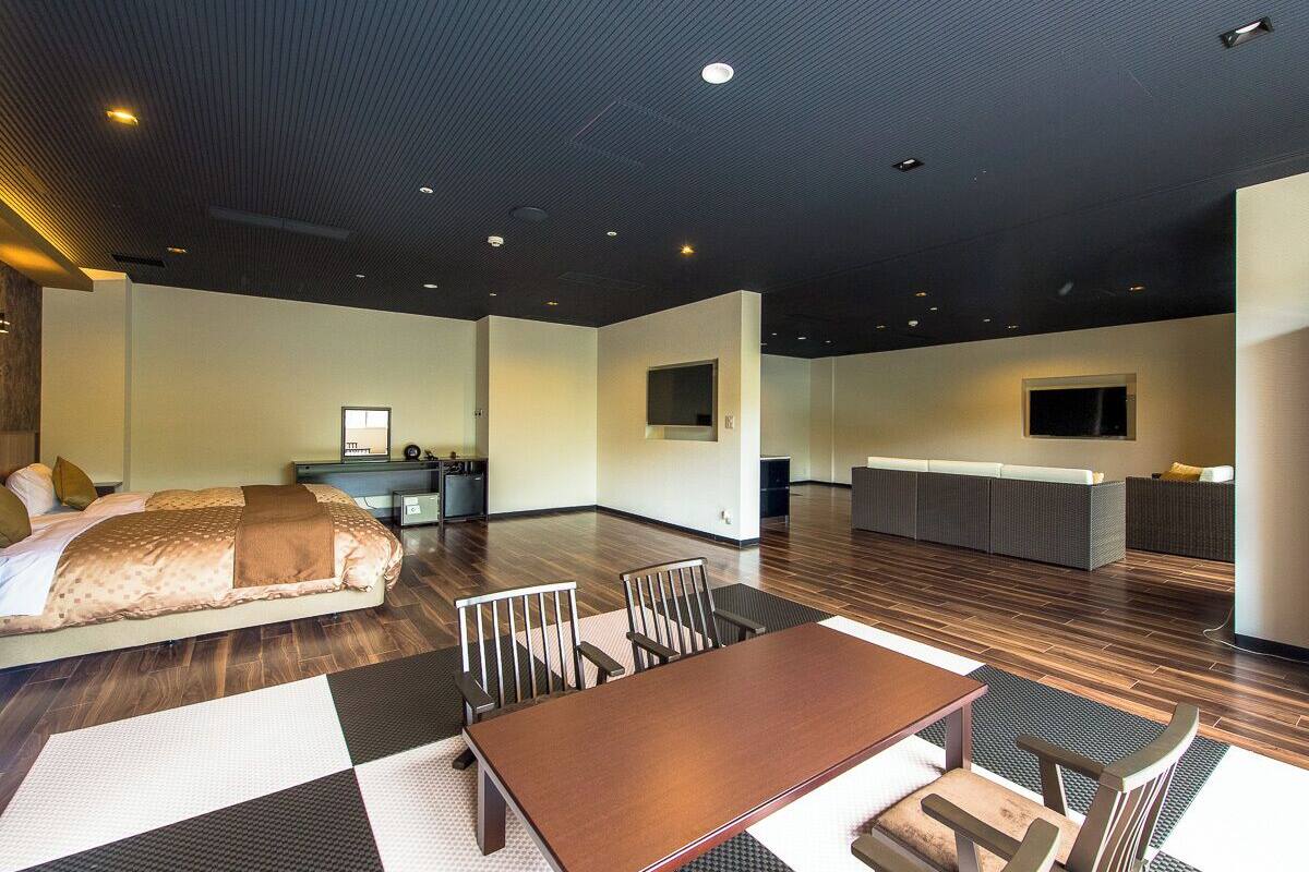 A modern hotel room with a large bed, a dining table, and a kitchen area.