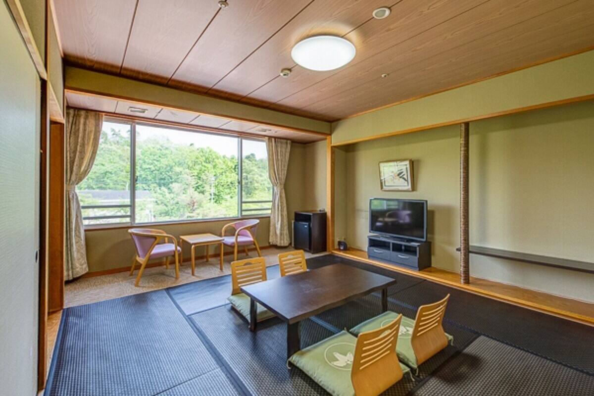 A room with tatami mats, a wooden table, chairs, a television, and a view of greenery outside.