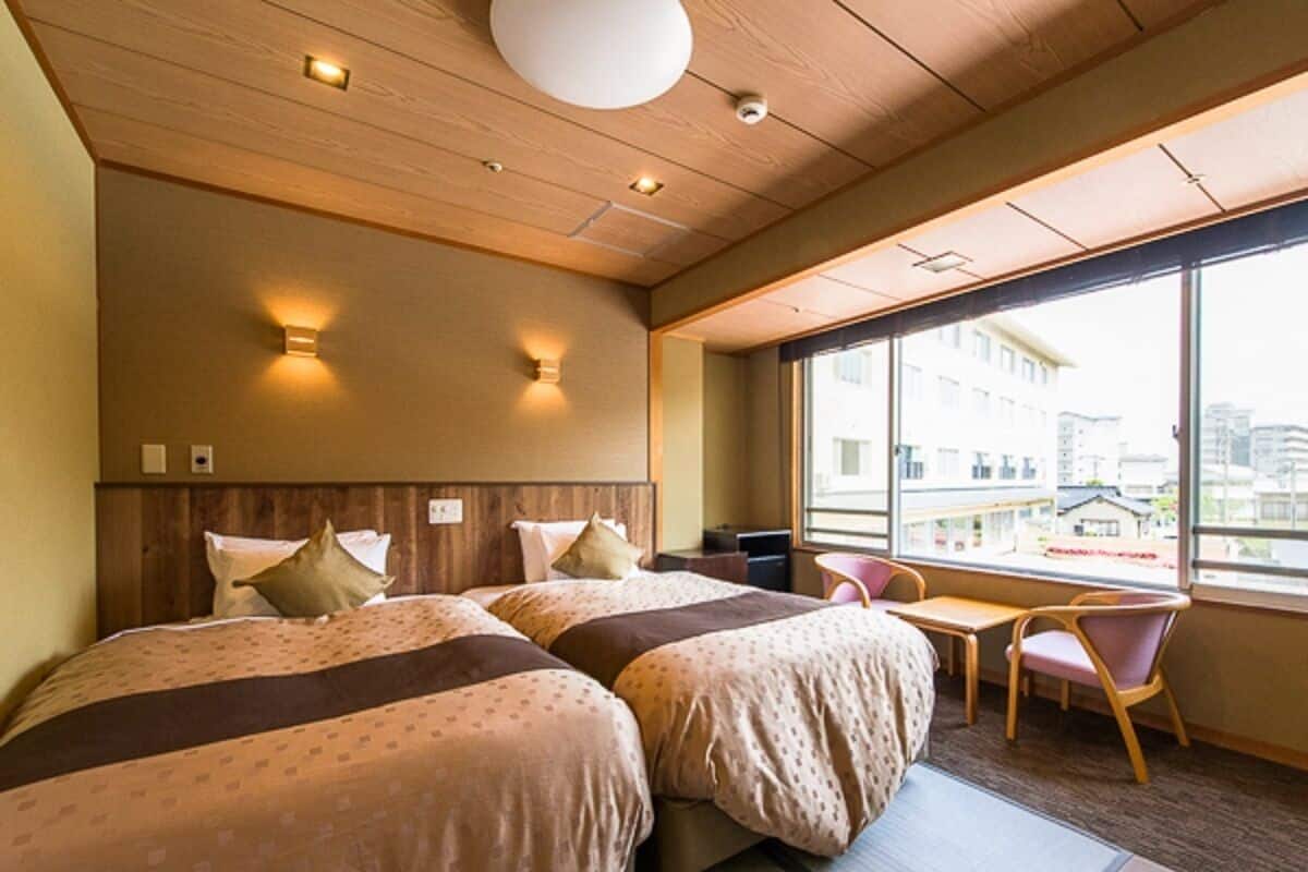 A hotel room with two beds, a desk, and a chair. There is a large window providing a view of buildings outside.