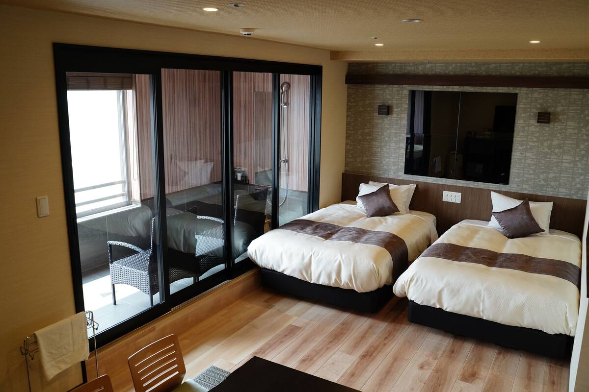 A modern hotel room with two beds, a large sliding glass door leading to a balcony, and a large mirror on the wall.