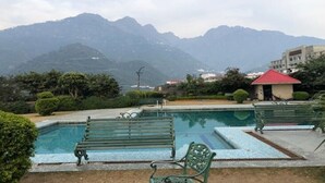 A swimming pool with benches, surrounded by greenery and mountains in the background. - Hotel Devi Grand (Reasi)