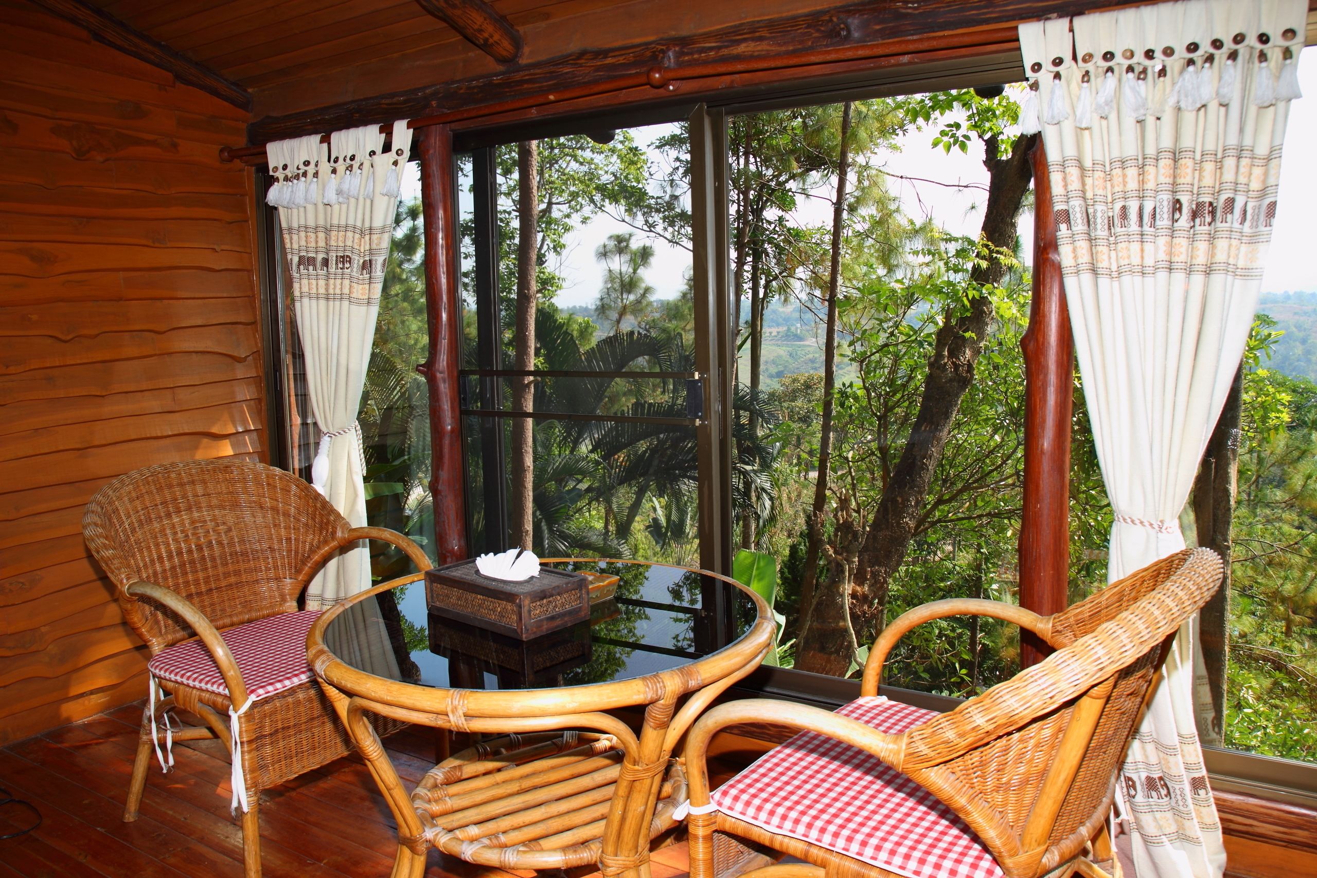 A wooden room with wicker chairs and a glass table, large windows with curtains, and a view of trees and hills.