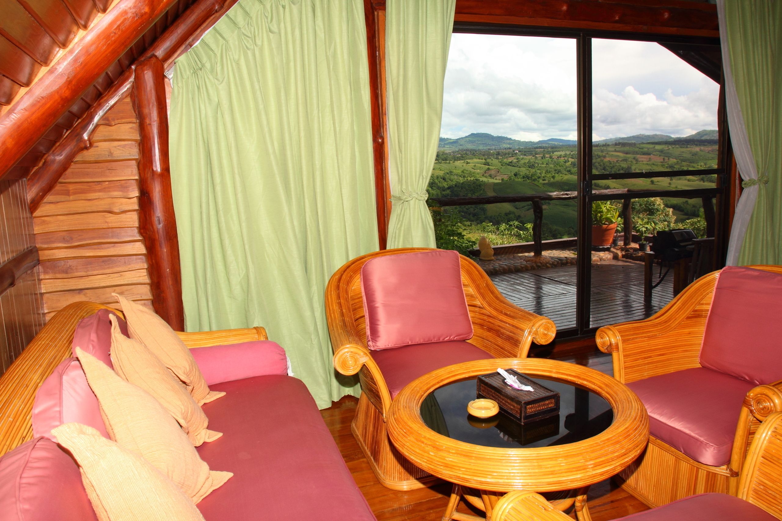 A wooden interior with a sofa, two armchairs, and a round table. A large window offers a view of green hills.