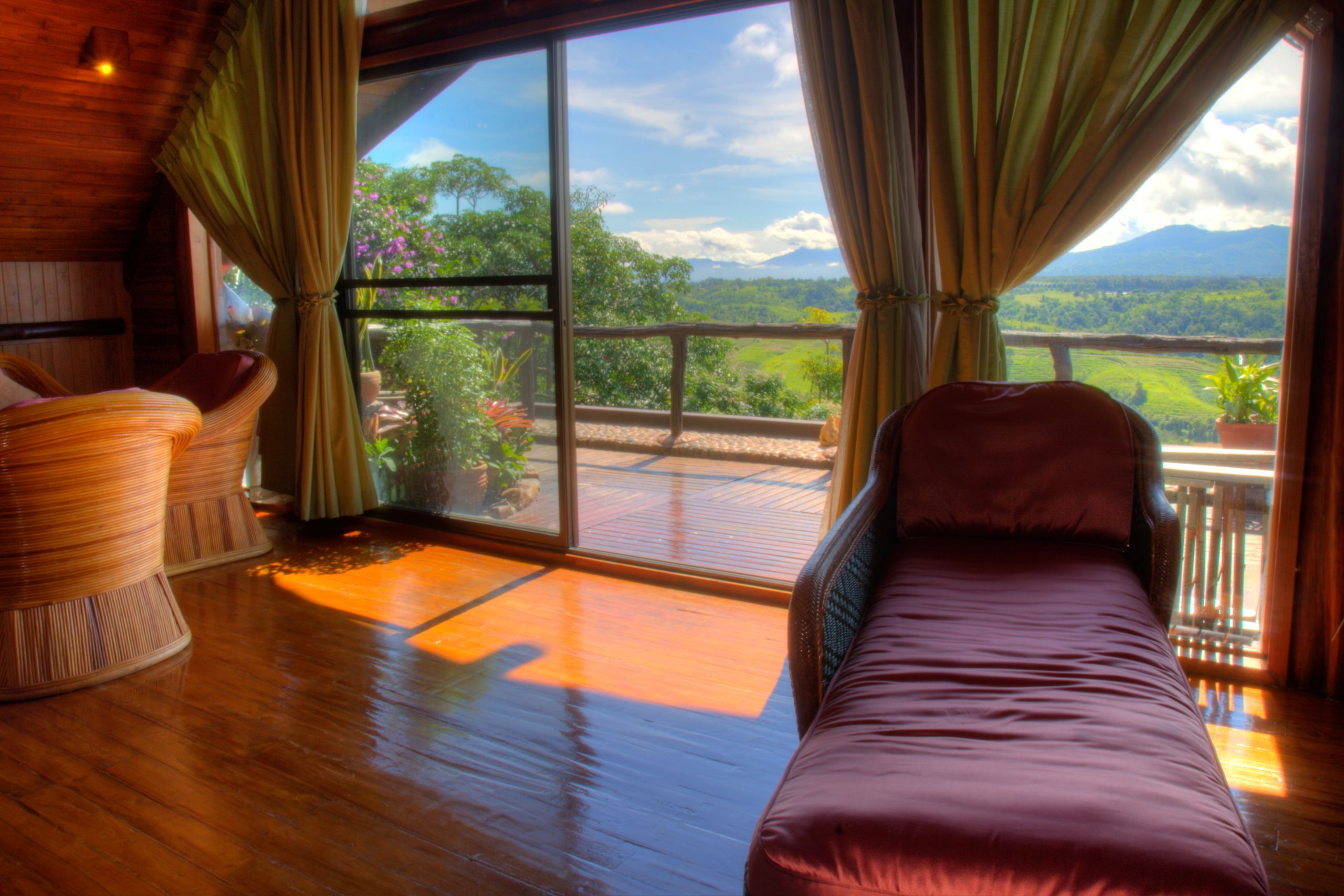 Habitación con un ventanal grande que ofrece vistas a un paisaje exuberante y montañas. Hay una cama y dos sillones de mimbre.