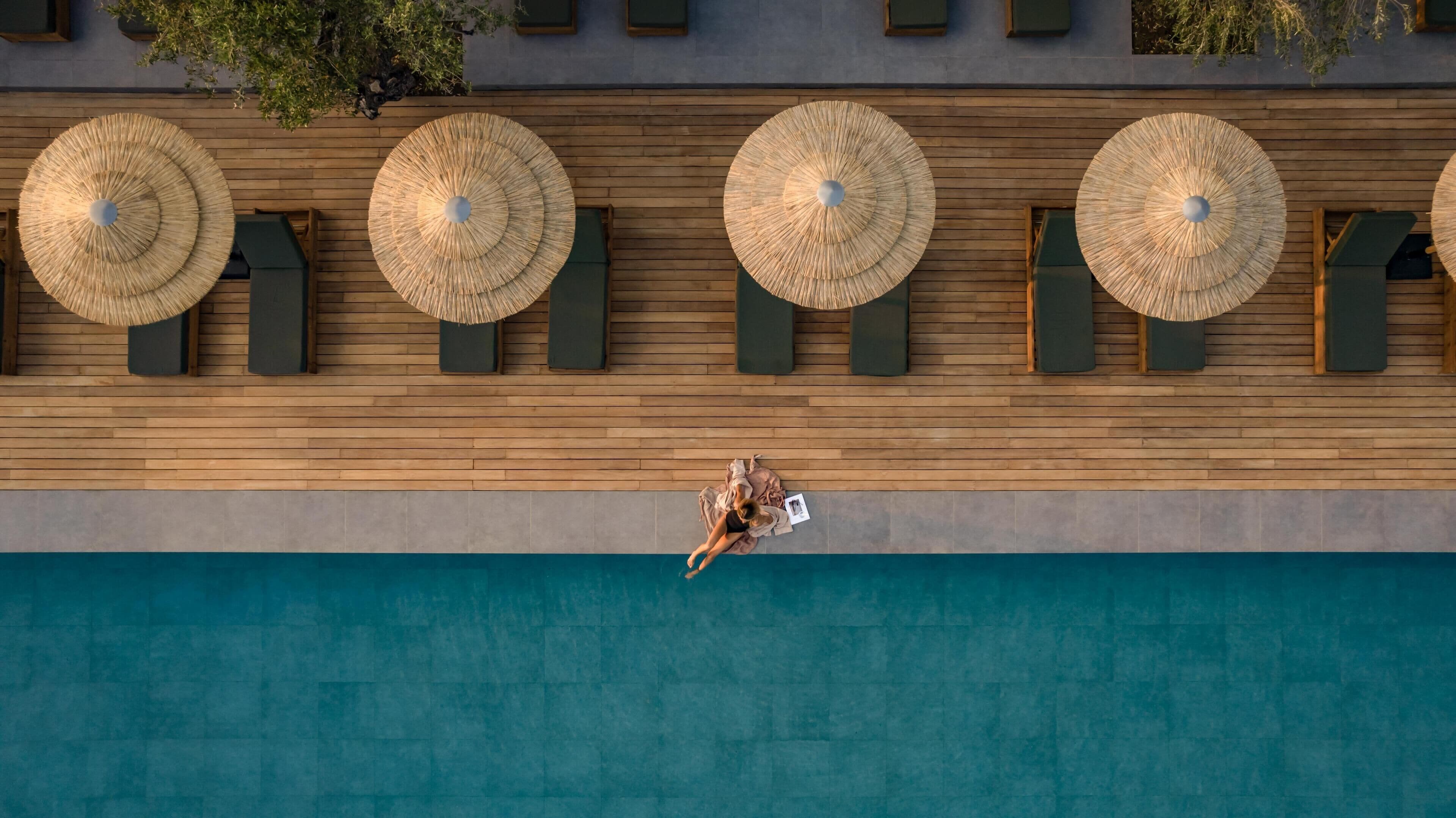 Vue aérienne d’un espace piscine agrémenté de quatre grands parasols en osier et de chaises longues disposées le long du bord de la piscine.