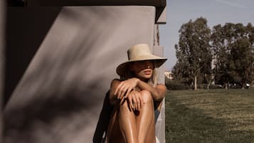 Una persona sentada en una silla con un sombrero grande, apoyada contra un edificio con un área verde y árboles al fondo.