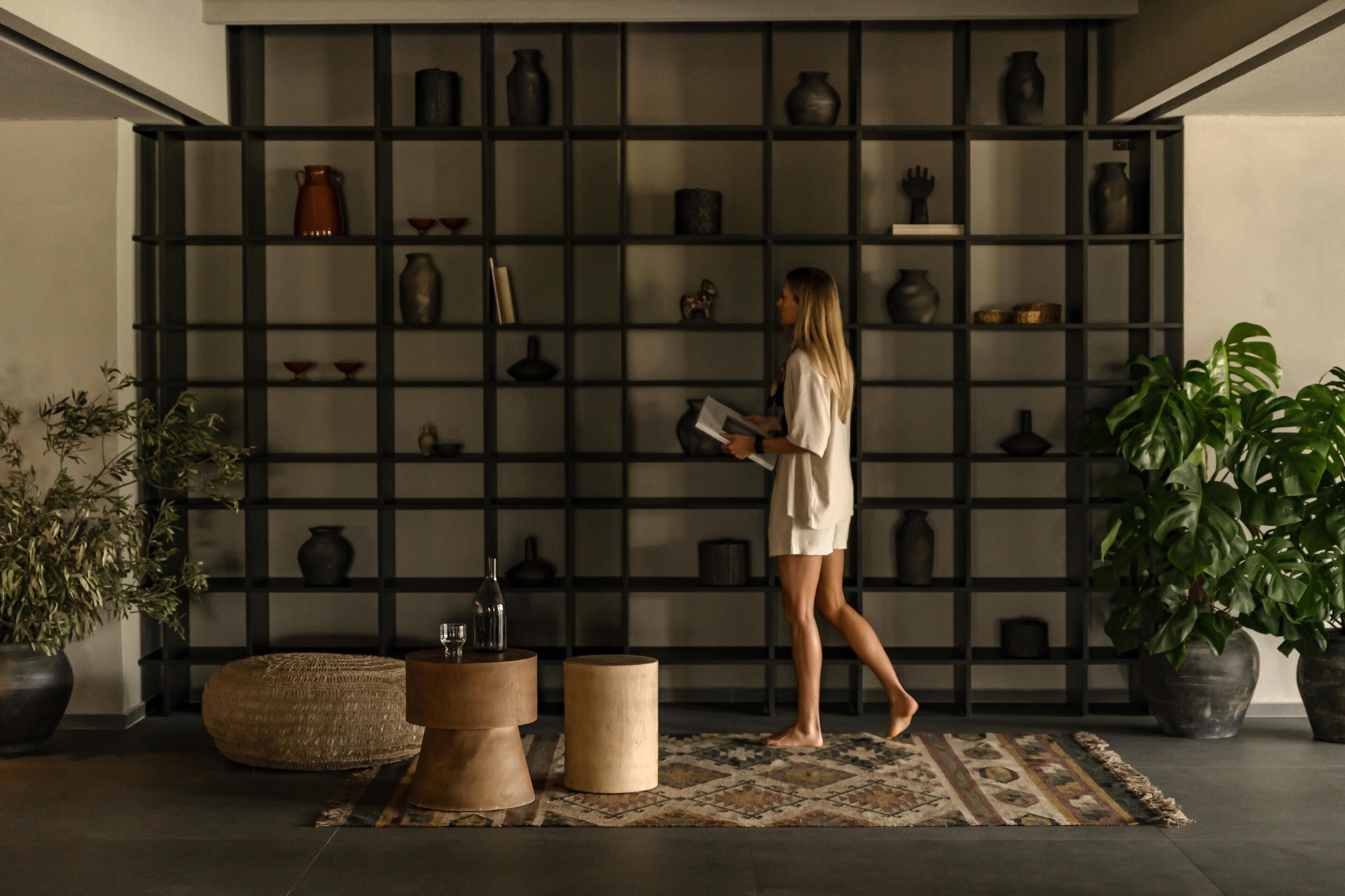 A room with a black shelving unit filled with various pottery and decorative items, a large potted plant, and a woven pouf.