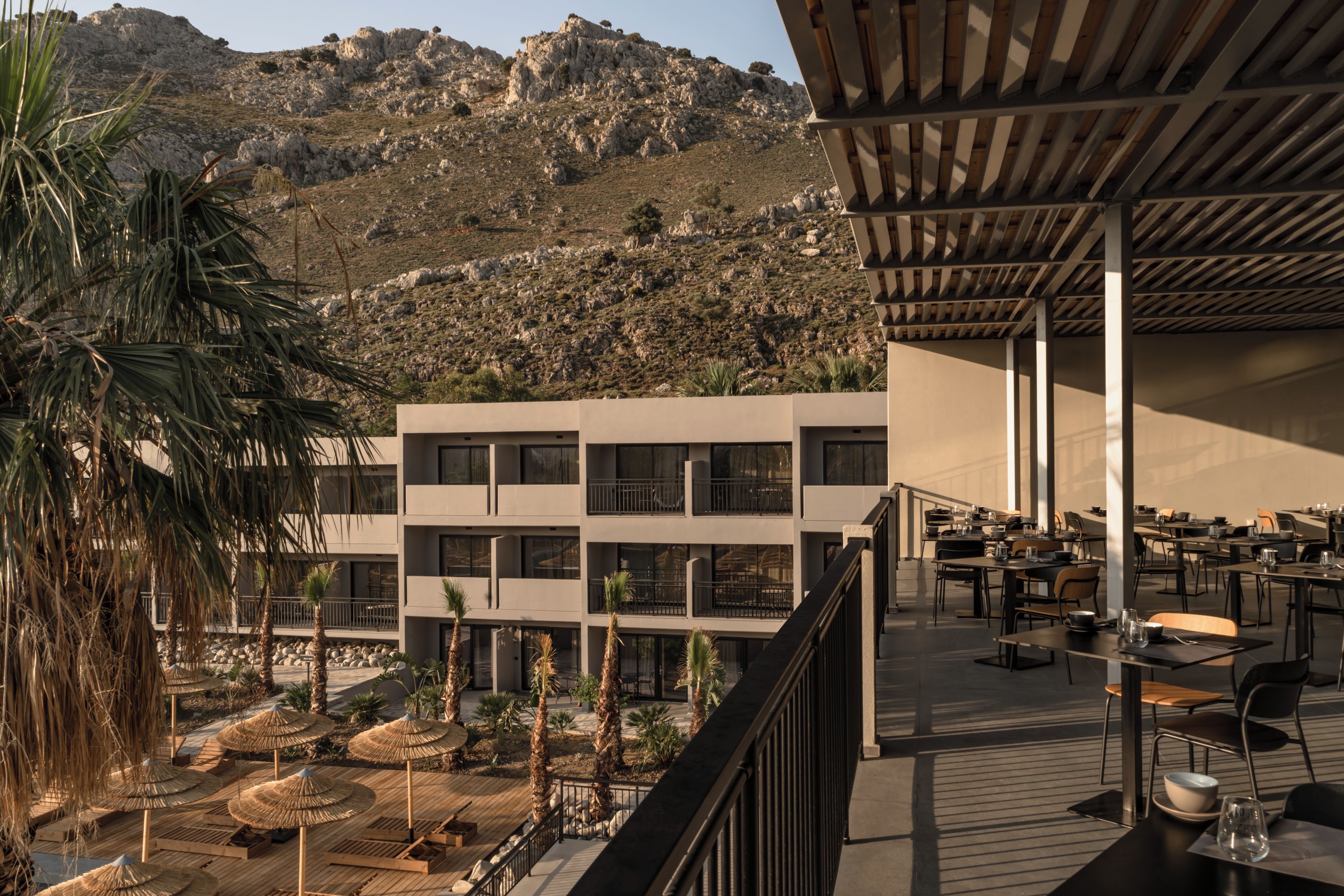 Un edificio moderno con varios pisos, una zona de estar al aire libre con sombrillas y vistas a colinas rocosas al fondo.
