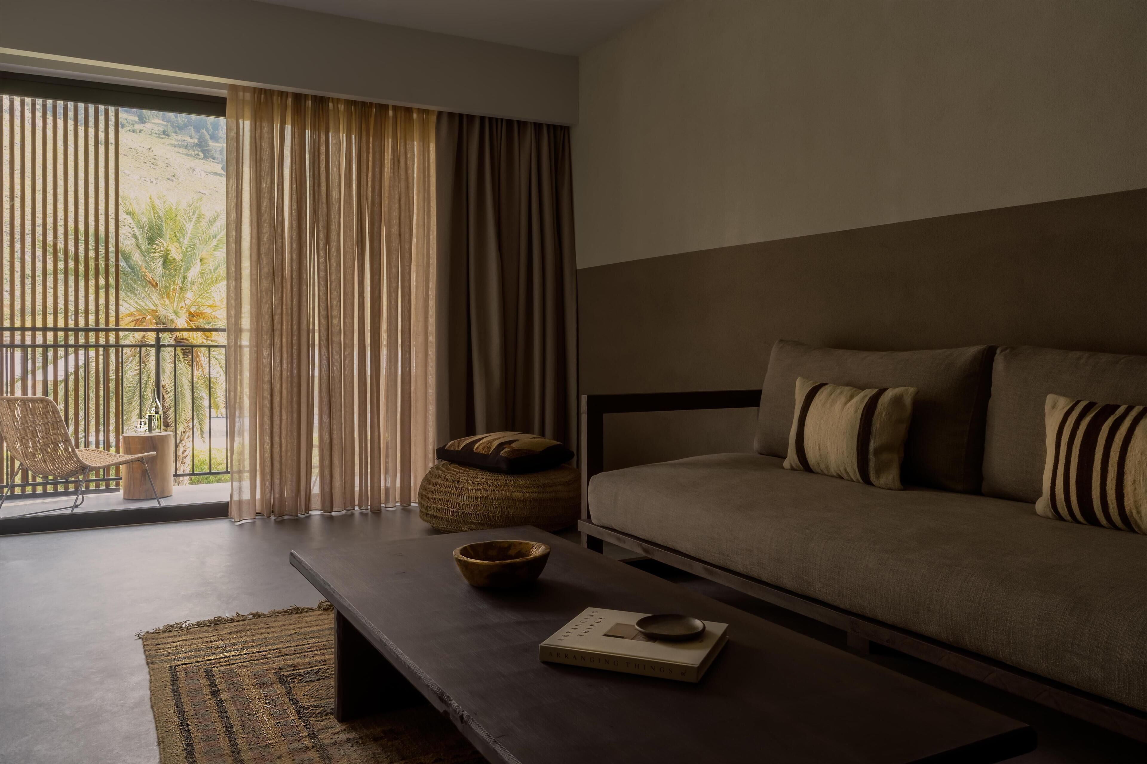 A modern bedroom with a large bed, a small side table, a chair, and a view of palm trees through the window.