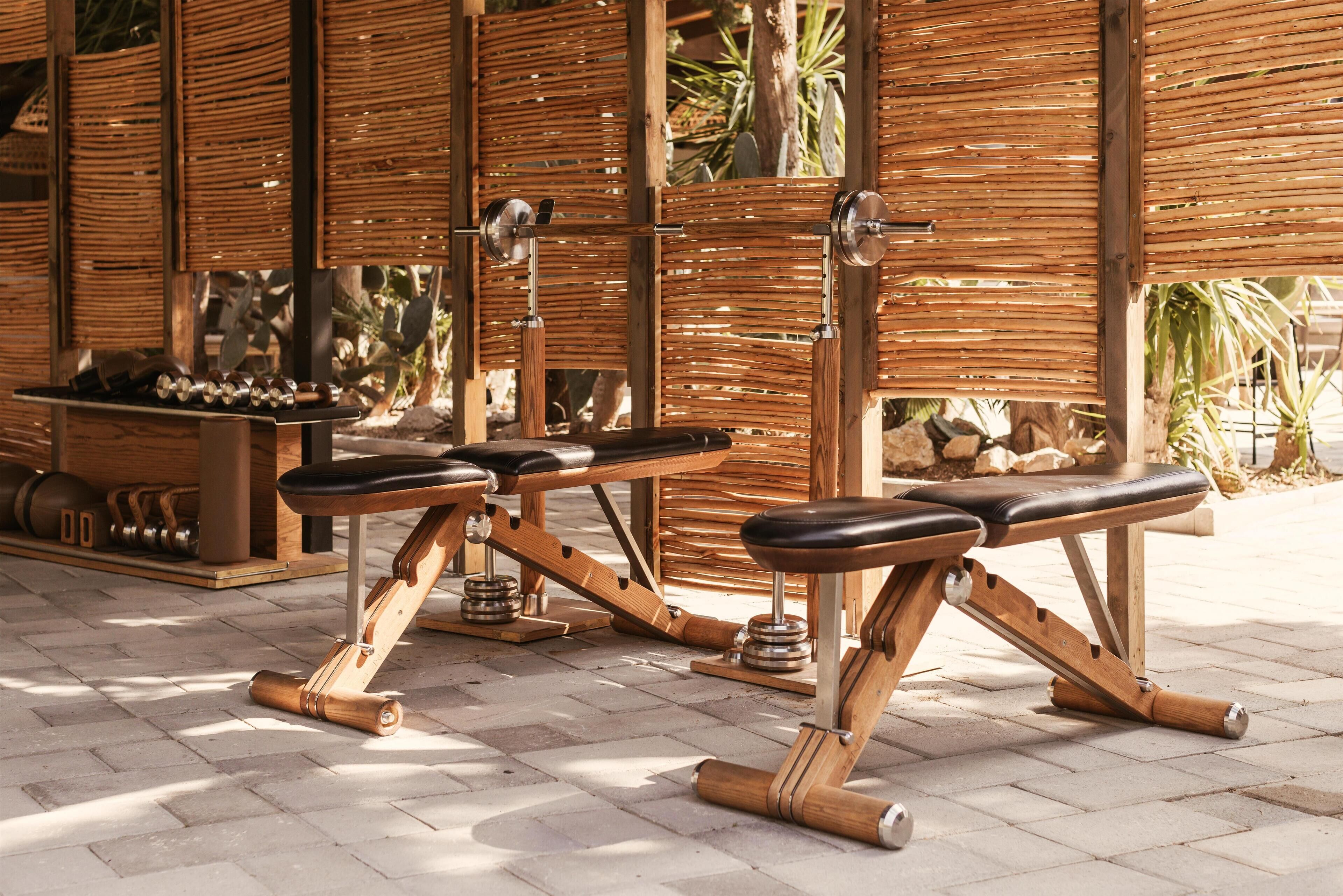 Outdoor exercise equipment, including benches and weights, in a patio area with a bamboo fence and potted plants.
