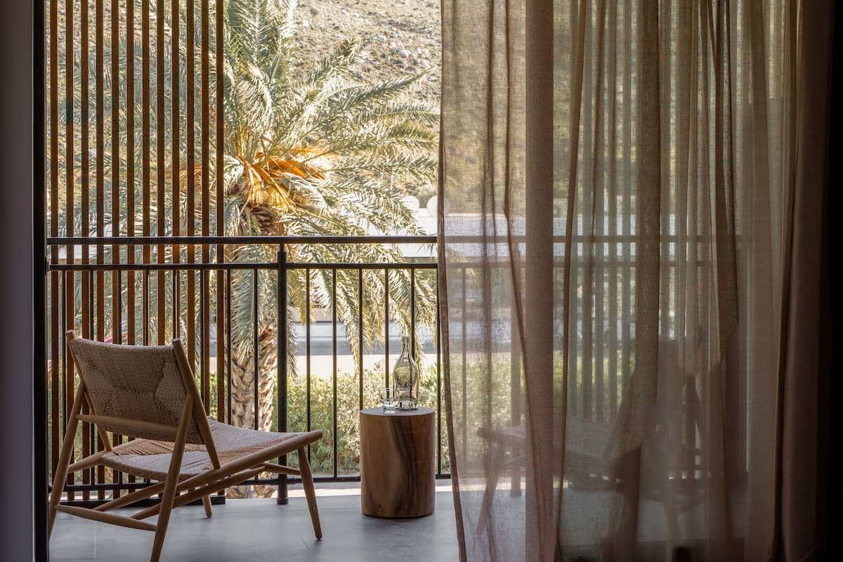 Un balcón con una silla tejida, una pequeña mesa de madera con un florero y vistas a palmeras a través de persianas verticales.