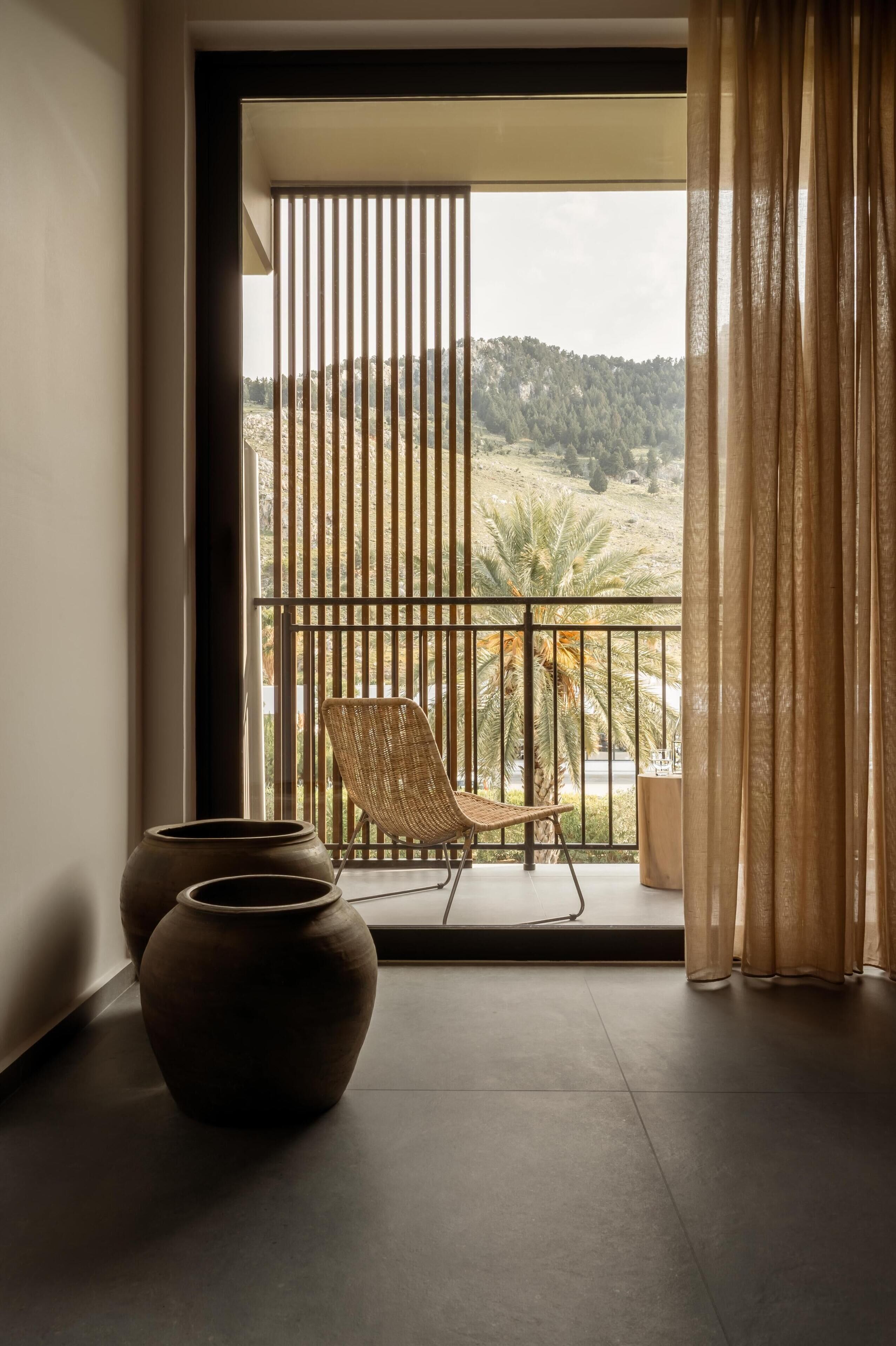 A balcony with a wicker chair and two vases, overlooking a landscape with trees and hills.